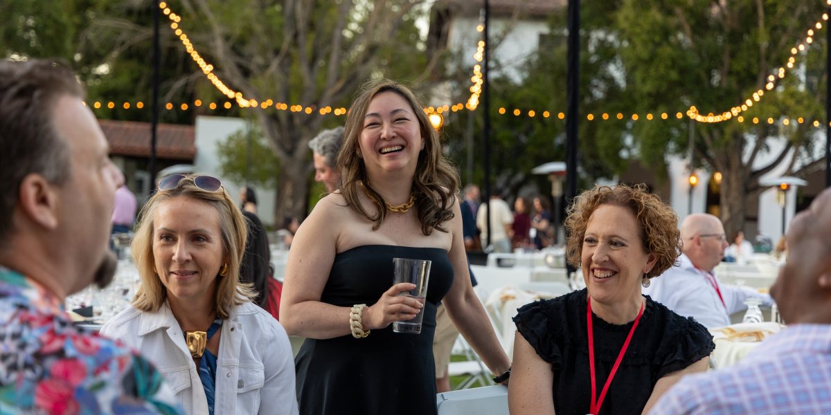 Five alumni laugh in conversation underneath string lights
