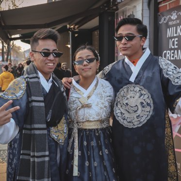3 students in traditional Chinese garb and pixel sunglasses pose for a picture on a busy street.
