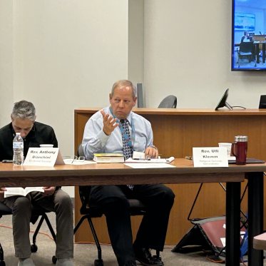 A person in shirt and tie, talks at a conference, with his name displayed on the table