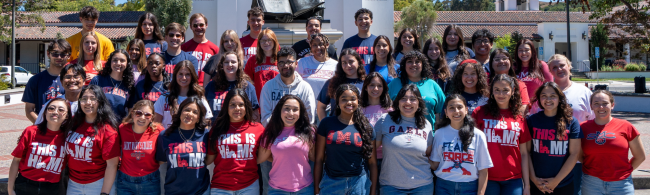 A group of students stand together outside, smiling for a group photo. Many are wearing school spirit shirts in red, blue, and white. A building and trees are visible in the background.