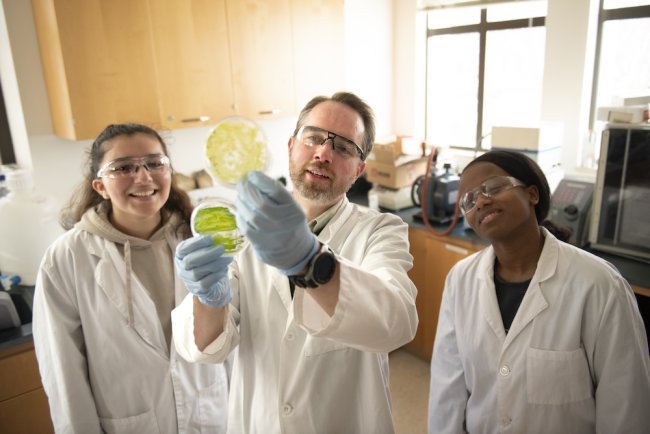 Prof. Jim Pesavento holds up Petri dish and two students in lab coats look at it