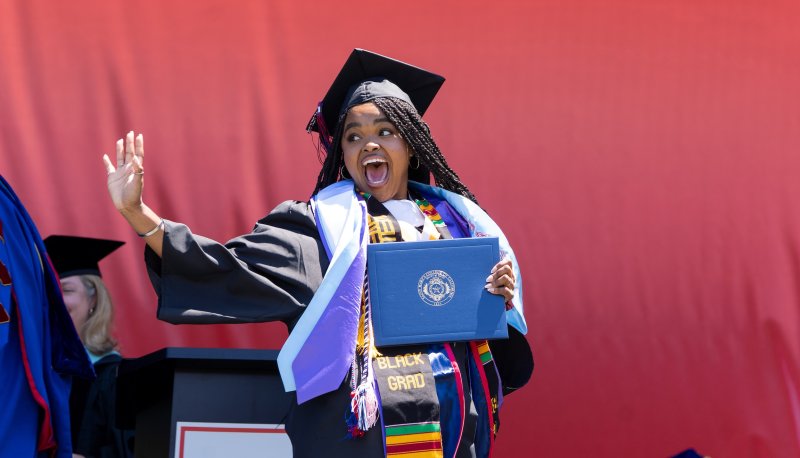 Female student holding diploma and smiling and waving at 2026 Undergraduate Commencement