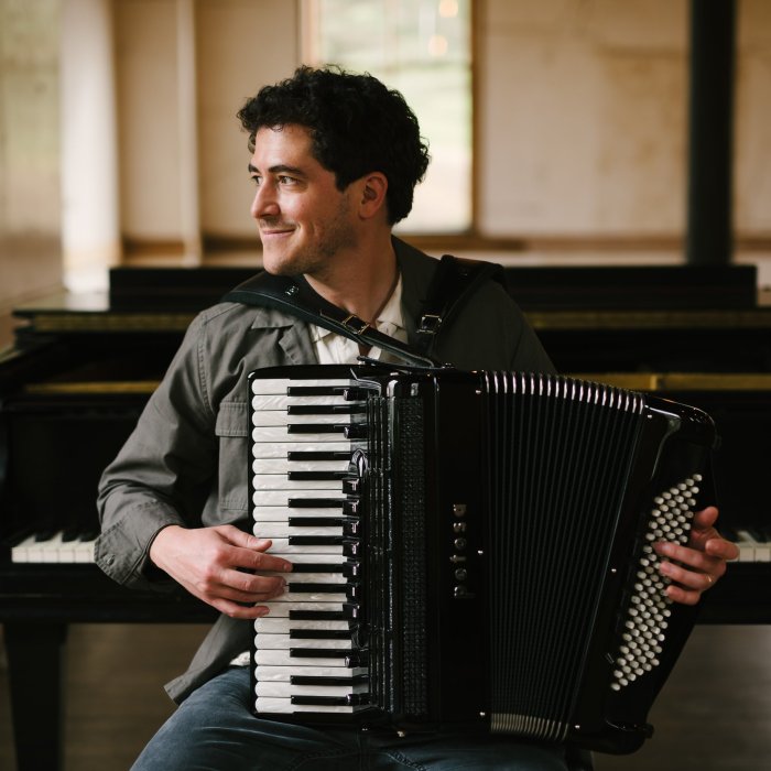 Sam Reider, lookng to the left, holding an accordion. Behind him, a piano.