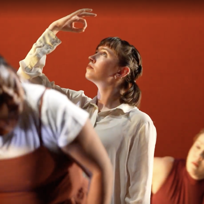 Three dances against a red background the one in the center is wearing a white button down shirt and is looking up at her raised hand