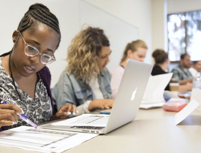 Professional MBA students sitting at a row of desks doing work on laptops and paper