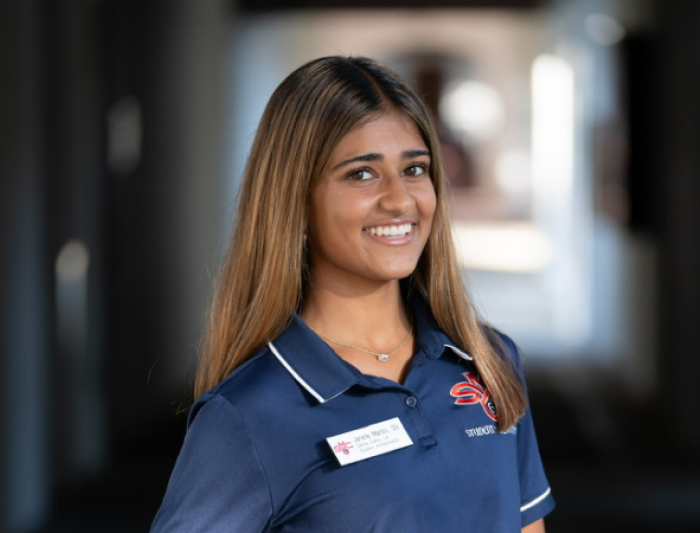 Portrait of a smiling student staff member wearing a navy polo shirt with a campus logo and name badge, standing in a softly blurred campus hallway.